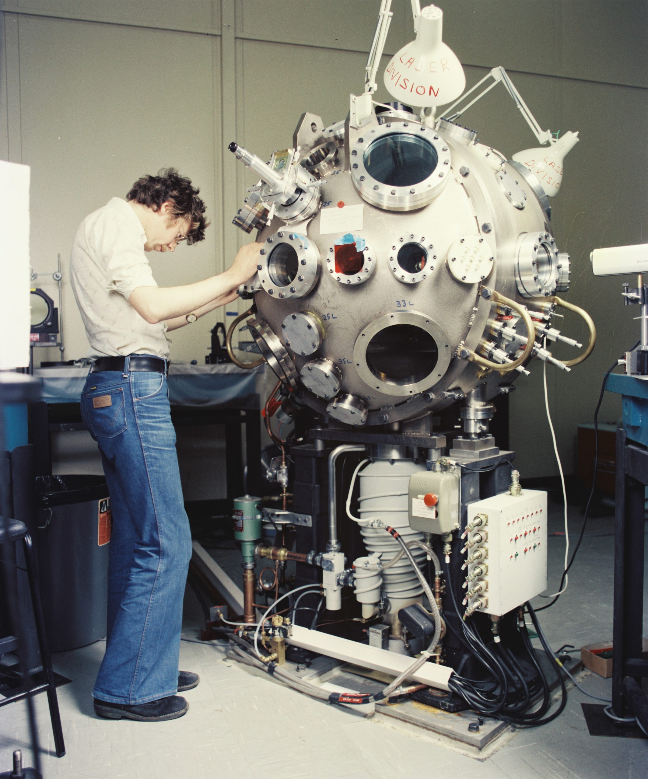 A person in 70’s attire stands working on a spherical metal chamber about the side of an exercise ball. The chamber is mounted onto a table and ha many round port holes.